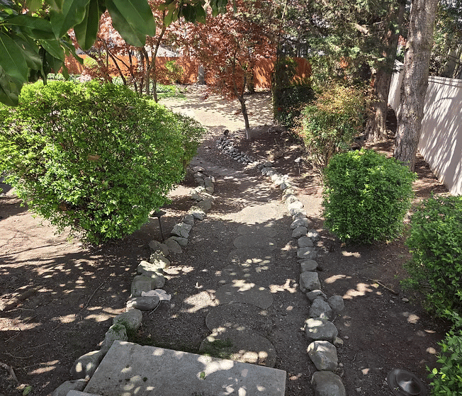Serene garden pathway lined with stones and lush green bushes leading to a tranquil space.